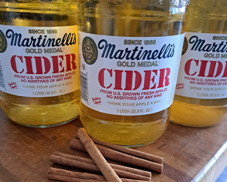 Three Martinelli's Gold Medal Apple Cider bottles arranged on a wooden cutting board, with several cinnamon sticks placed in front. The center bottle is in sharp focus, while the bottles on the left and right are partially cropped.