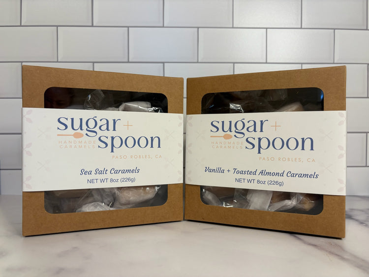 Two packages of 'Sugar + Spoon' caramels on a marble surface with a tiled wall background.